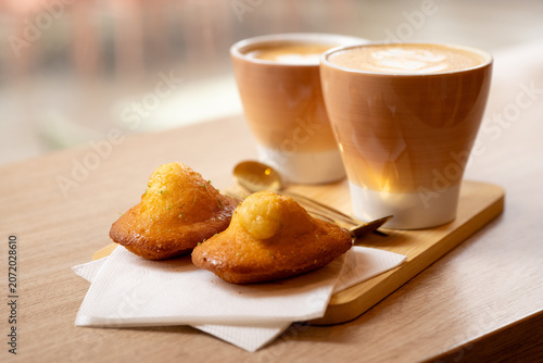 Foto Two Artisan Lattes with Latte Art Served with French Madeleines on Wooden Board - A beautifully styled café scene featuring two gradient ceramic cups of latte with intricate milk foam art