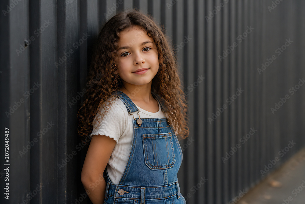 Fototapeta premium A young girl with curly brown hair wearing denim overalls stands against a dark corrugated metal fence.