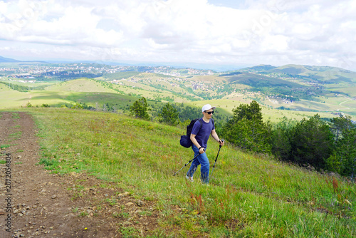 Принти на полотні Man with trekking poles follows open upland route across panoramic terrain overlooking valley with resort town