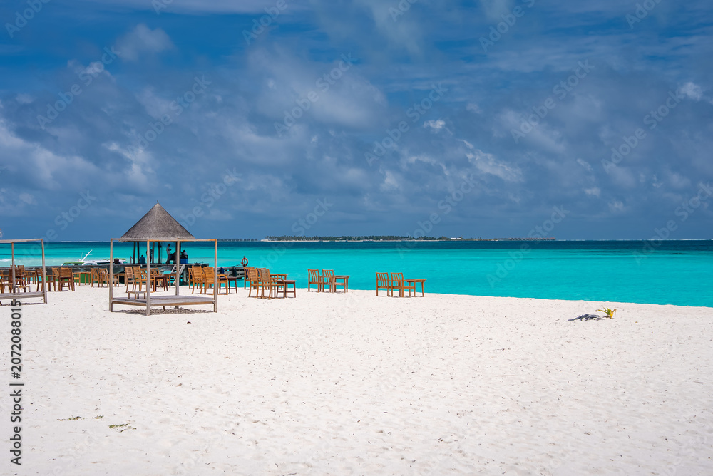 Fototapeta premium White sand stretches to turquoise water with lounge chairs and a thatched pavilion in the Maldives. Soft daylight and wide view suggest a quiet resort setting.