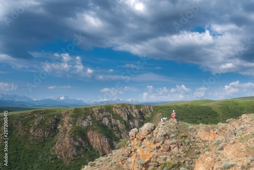 Woman with dog sits on a rocky cliff edge overlooking a vast canyon valley under a cloudy sky. Multi-purpose lifestyle stock photo for travel, outdoor adventure, freedom concept, or banner background.