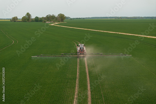 Canvas Print A wide boom sprayer moves along parallel tire tracks, misting fine droplets over uniform young crops, a pale dirt farm road curving in distance under soft overcast light
