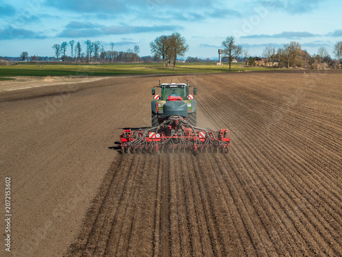 A modern tractor pulls a multi row seed drill, carving straight furrows across a wide tilled field under cool daylight, leafless trees and a windbreak line visible.