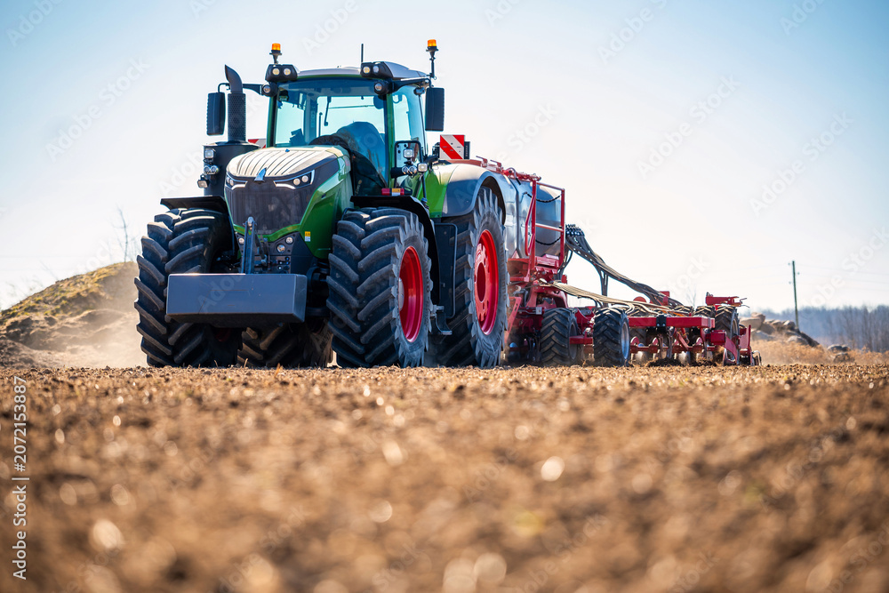 Fototapeta premium Modern Fendt tractor with oversized tires tows a red multi row seeder across tilled soil, dust rises, GPS guided planting in spring, low angle view shows tread and hoses.