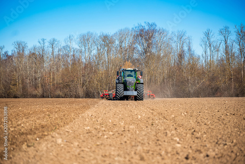 Obraz na plátně A green tractor with dual lights pulls red tillage toward the viewer on a freshly plowed field, dust rising, bare deciduous treeline in bright daylight, cool season