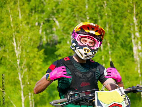 Man enduro rider in protective helmet and gear sitting on motorcycle in forest. Motocross racer showing gesture of success. Extreme sport athlete enjoys adventure and outdoor hobby.