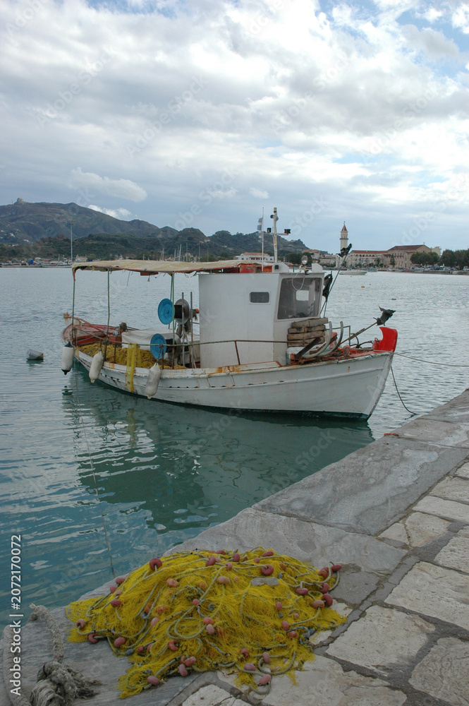 Fototapeta premium A traditional white wooden fishing boat moored at a stone dock with a pile of yellow fishing nets in Zakynthos port under a cloudy sky