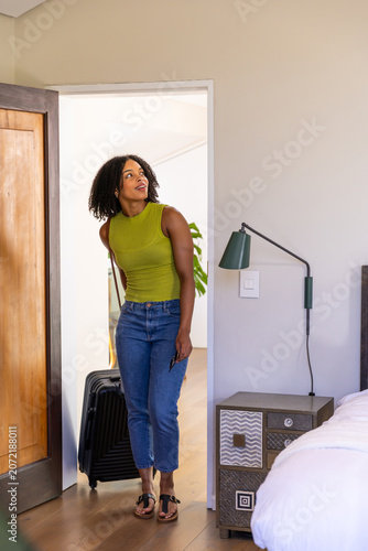 African American woman standing at bedroom door pulling suitcase in green top, jeans holding phone