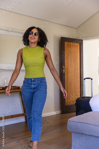 Adult African American woman walking through bedroom wearing lime top, sunglasses, suitcase nearby