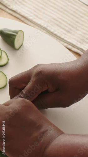 Vertical video: Starting holding blue knife, hands slicing cucumber on cutting board for white bowl