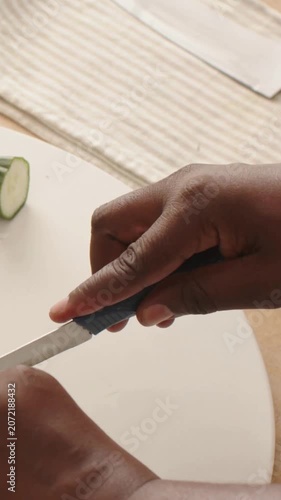 Vertical video: Picking up blue knife, adult hands slicing cucumber into rounds on board for salad
