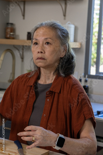 Senior Asian woman sitting at countertop preparing veg on cutting board with blue knife smartwatch