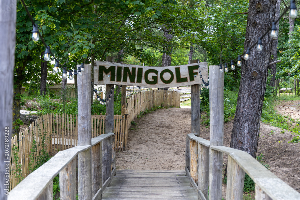 Obraz premium Wooden bridge leading to a minigolf course entrance surrounded by trees and a sandy path, with decorative lights strung overhead for ambiance