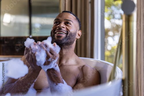Valokuva Mature man sitting bare-chested in freestanding white bathtub holding suds and s