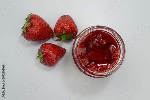 Homemade strawberry jam in glass jar with ripe strawberries arranged on white background. Fresh berry preserve concept.