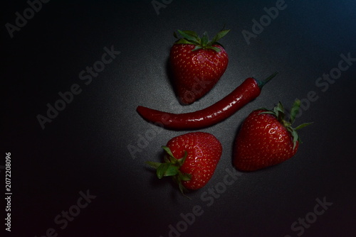 Creative minimalist arrangement of strawberries and red chili pepper on dark background. Sweet and spicy flavor concept with copy space.