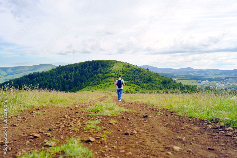Obraz premium Small figure walking through wide upland scenery with winding route stretching toward forested hills in the distance. Conveys landscape-scale recreation and exploration of expansive environments.