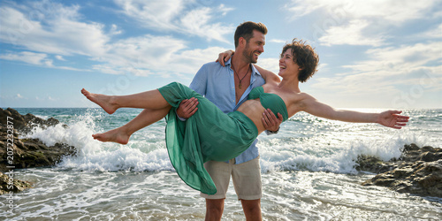 Man in blue shirt holding smiling woman in green dress embracing on a rocky beach with ocean waves and blue skies with white clouds in the background.