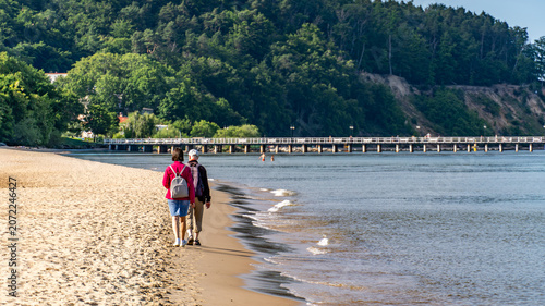 Senior Couple Walking Along Baltic Sea Beach with Backpacks on Sunny Day