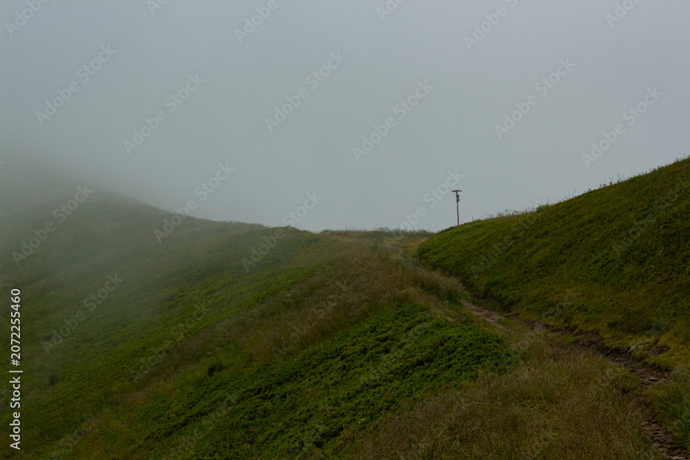 Obraz premium Narrow dirt path winding up a vivid green mountain hill disappearing into low clouds and dense fog. Minimalist moody landscape capturing pure solitude, mystery and the call of wild highland adventure.