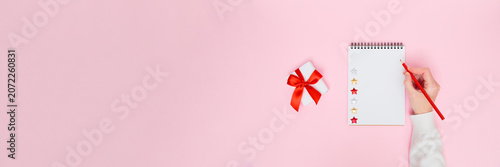 Female hand writing on blank notepad with gift box and red ribbon on pastel pink background, festive flat lay banner with copy space for holiday promotion, planning, goals, to-do list or wish list