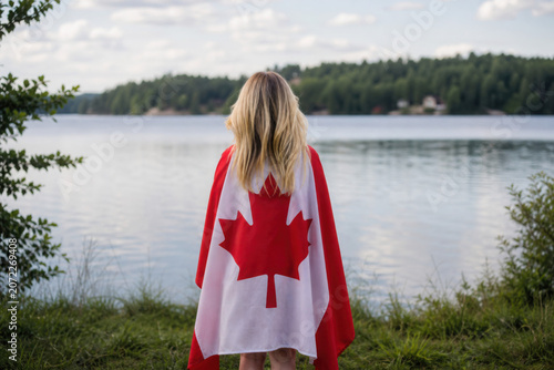 Girl wrapped in large Canadian flag by Muskoka lake in nature. Canada Day celebration outdoors. Kid in large Canadian flag celebrating national Canada Day on 1 of July.