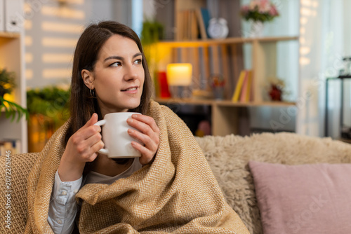 Quadro su tela Young woman on home sofa holds hot drink wrapped in blanket warming up and enjoying quiet rest at home