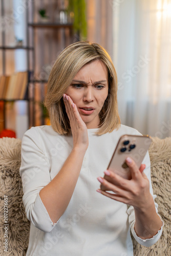 Obraz na płótnie Middle-aged sad woman sits on home sofa after reading bad news on smartphone, sighing in disappointment
