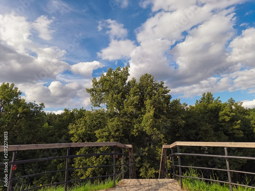 Wood fence on a view of green park with trees and blue sky with beautiful fluffy clouds in sun summer day. Natural nature cloudscape bright background