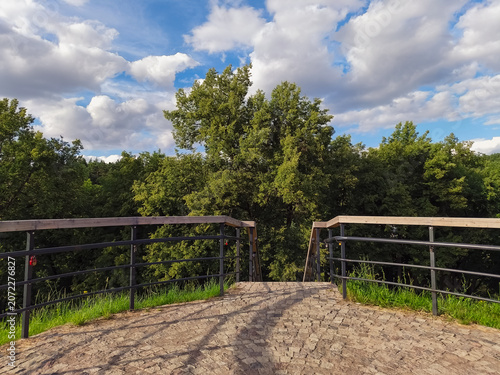 Wood fence on a view of green park with trees and blue sky with beautiful fluffy clouds in sun summer day. Natural nature cloudscape bright background