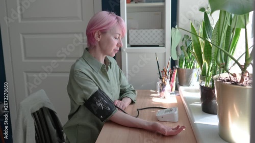 Mature woman seated at table preparing to check blood pressure using tonometer, female taking care of health at age of 50, using electronic device monitoring results. Reaction to change in weather 