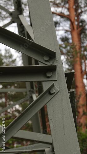 Zdjęcie przedstawia zbliżenie na metalowe kątowniki i śruby tworzące stalową konstrukcję kratownicową, z rozmyciem pni drzew w tle.