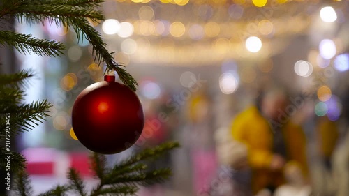 Red christmas ball ornament hanging on a fir tree branch. Festive red christmas ball ornament hanging on a fir tree branch with a beautifully blurred background of city lights and people walking