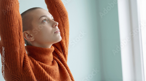 A pensive young woman with cancer and a shaved head wearing an orange turtleneck sweater gazes upward, symbolizing hope and resilience in a bright indoor setting with soft natural light.
