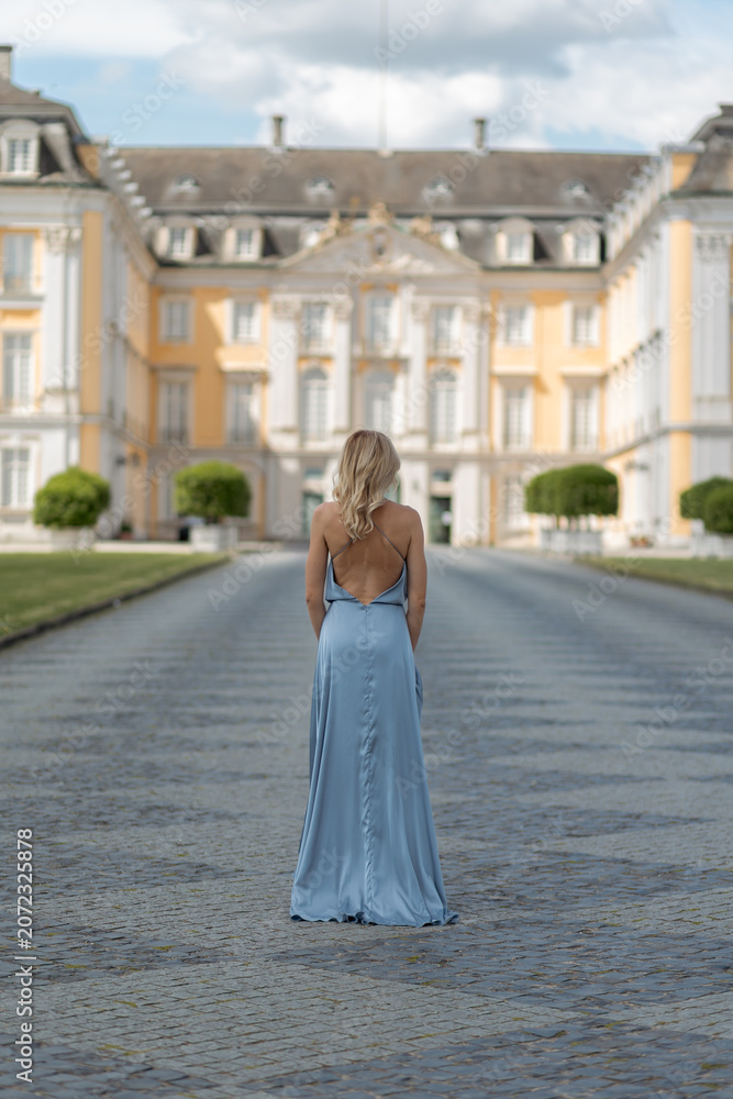 Obraz premium Woman walks alone on stone path in front of large historic building with yellow walls and manicured lawns in afternoon light