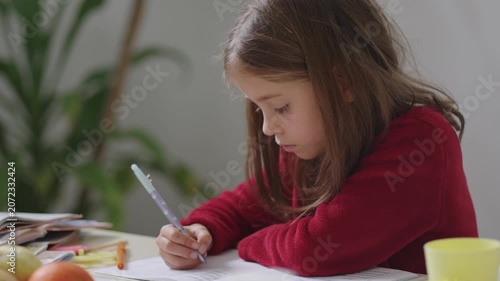 A determined young girl sits at her desk, concentrating on her homework. Surrounded by vibrant fruits and colorful stationery, she writes with focus, creating art with words.