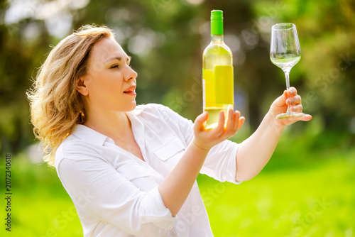 Φωτογραφία Blonde woman looking at white wine bottle and holding empty clear wineglass outdoors