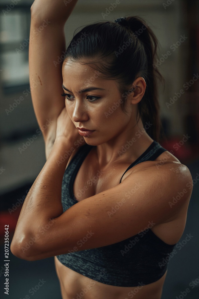 Fototapeta premium Fit woman stretching arm overhead in gym, wearing sports bra and glistening with sweat, symbolizing strength, motivation, healthy lifestyle, and modern fitness culture.