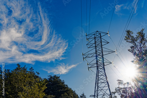 Looking at an electricity pylon and transmission power lines with the sun behind and blue sky with swirly white clouds above in Arthur's Pass, New Zealand.