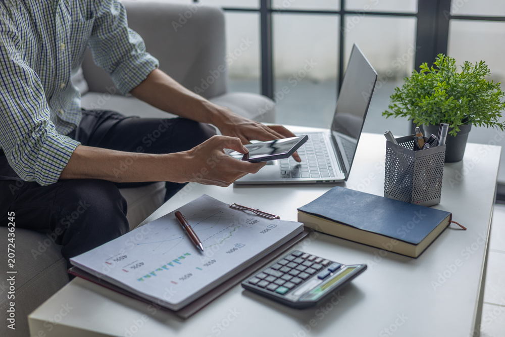 Fototapeta premium Businessman calculating expenses and reviewing financial statistics using smartphone and laptop.