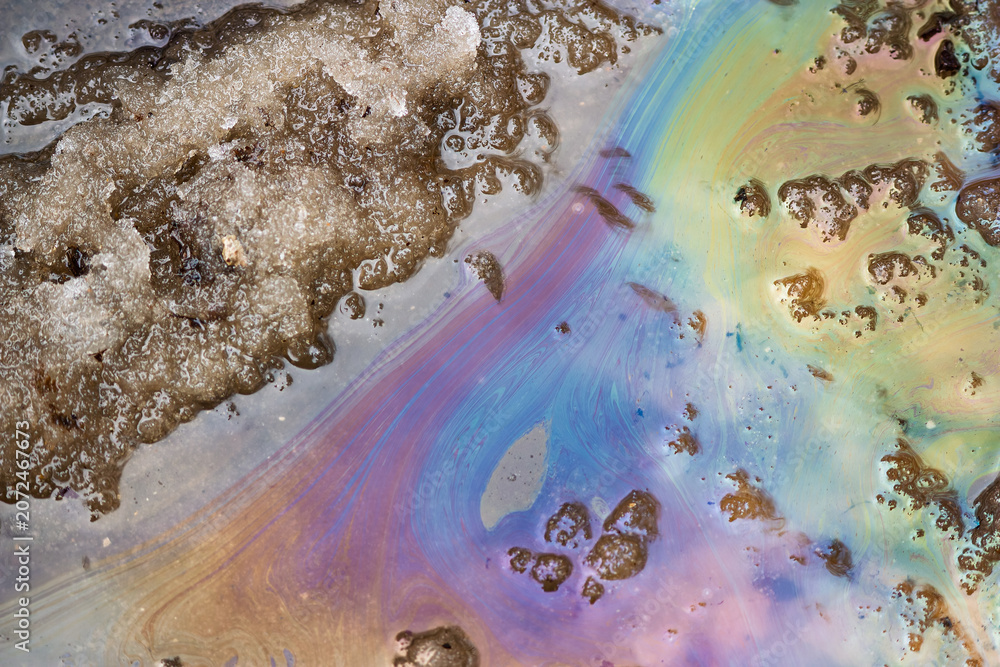 Naklejka premium Iridescent oil slick on a puddle surface with melting dirty snow, showing colorful petroleum spill abstract patterns after winter thaw on an asphalt road in urban environment