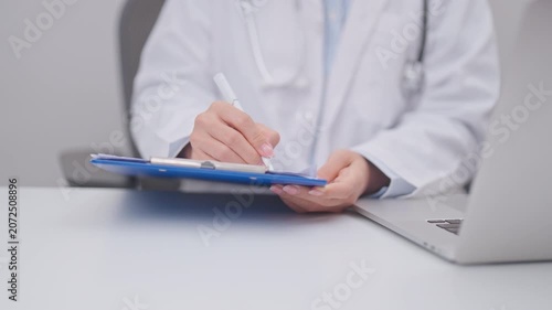 Professional doctor wearing lab coat and stethoscope using laptop computer and taking report notes on notebook at desk in hospital clinic office, medical technology and healthcare concept