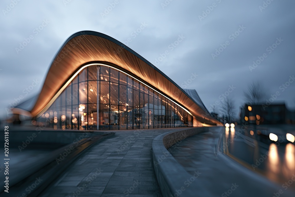 Fototapeta premium Modern architectural structure with curved, illuminated roof and glass facade at dusk