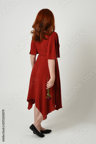 Full length portrait of beautiful redhead model wearing elegant red modern dress. standing pose, walking away facing backwards, holding a romantic red rose, isolated figure studio background