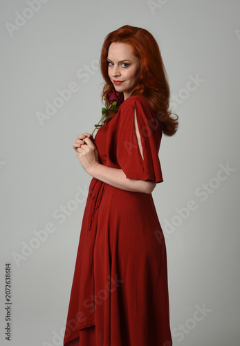 close up portrait of beautiful model with long red hair, wearing an elegant red modern dress. standing pose holding a romantic red rose, isolated figure on studio background.
