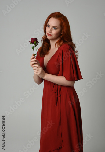close up portrait of beautiful model with long red hair, wearing an elegant red modern dress. standing pose holding a romantic red rose, isolated figure on studio background.
