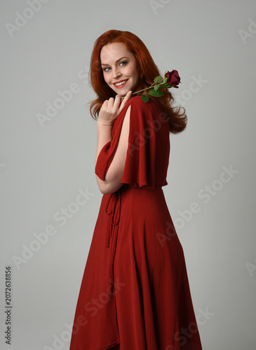 close up portrait of beautiful model with long red hair, wearing an elegant red modern dress. standing pose holding a romantic red rose, isolated figure on studio background.

