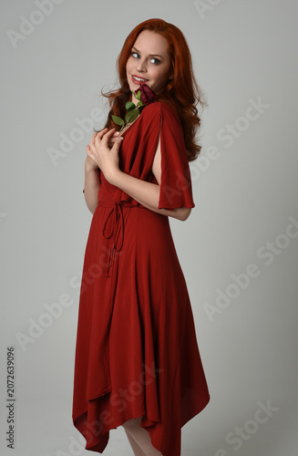 close up portrait of beautiful model with long red hair, wearing an elegant red modern dress. standing pose holding a romantic red rose, isolated figure on studio background.
