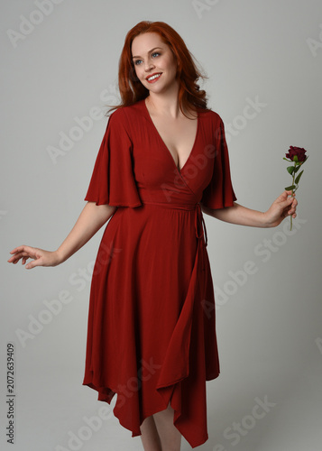close up portrait of beautiful model with long red hair, wearing an elegant red modern dress. standing pose holding a romantic red rose, isolated figure on studio background.
