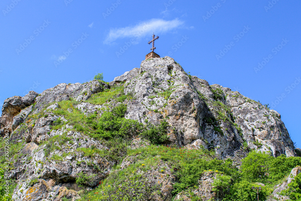 Fototapeta premium A tall cross on a rock covered with bushes and grass. Blue sky with clouds. Elia, Kakheti, Georgia